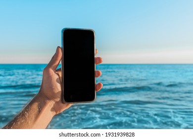 The man's hand holds a mobile phone with a black screen space for text and design against the blue sea, sky and skyline. - Powered by Shutterstock