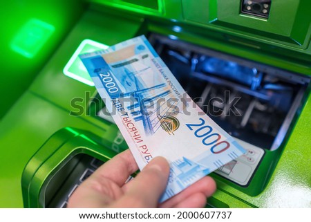 Man's hand holds new Russian banknote of two thousand rubles against background of green cash machine