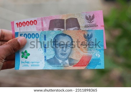 A man's hand holds a rupiah banknote against a blurred background. 100000 Rupiah and 50000 Rupiah.