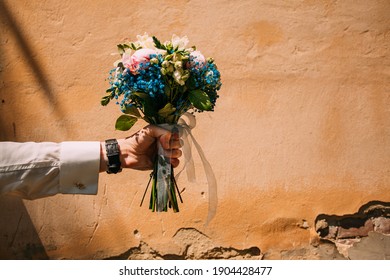 a man's hand holds a wedding bouquet of blue flowers against a background of orange grunge wall