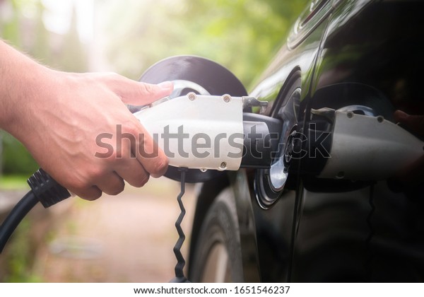 Mans hand\
inserting charger plug into electric car in green environment\
background. New energy vehicle, NEV is being loaded with\
electricity power. Ecology, modern day\
cars