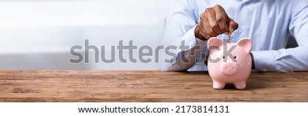 Man's Hand Inserting Coin In Piggybank Over Wooden Desk