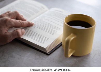 Man's hand on the book and a yellow cup of coffee on the table. Man is reading a book. Close up view. - Powered by Shutterstock