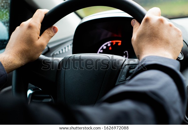 man\'s hand on steering wheel, driving a car\
Selective focus / close up from a man\'s hand on a steering wheel in\
a rainy day\
