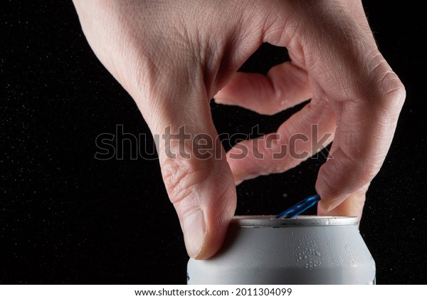 A man\'s hand opening a soda\
can on a black background and showing the splash when opening the\
can.