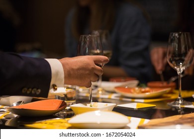 Man's Hand Picks Up Glass Of Wine At Fancy Dinner In Restaurant. Black Suite With Surgeon Cuffs, Arm Only, No Face. Fine Dining, Elegant People. Business Meeting Concept.