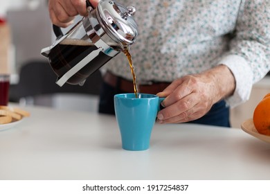 Man's hand pouring aromatic coffee into a blue cup from french press on a wooden table during breakfast. Elderly person in the morning drinking fresh brown espresso from vintage mug, filter relax