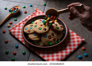 Man's hand pouring chocolate chips with wooden spoon onto vanilla cookies with red, blue, green and yellow chocolate chips on a black round plate on a red napkin with white checks, surrounded by choco - Powered by Shutterstock