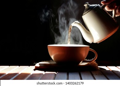 Man's hand pouring hot tea from ceramic teapot into tea cup with steam on wooden table in dark vintage tone style