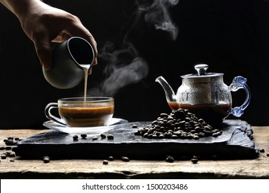 Man's hand pouring milk into a glass of espresso coffee with steam on black stone plate with wooden table in vintage dark tone style