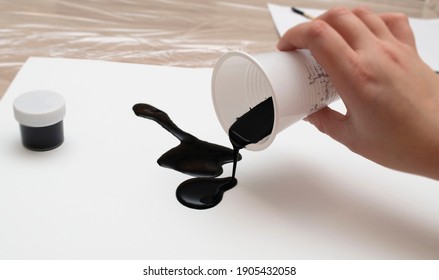 A Man's Hand Pours The Mixed Paint Onto A White Sheet Of Paper. Next To It Is A Closed Jar Of Black Acrylic Paint