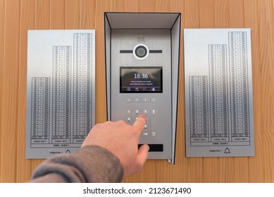 A man's hand pushes the buttons of a intercom with a camera and panels indicating the numbers of each floor.On the display you can read 