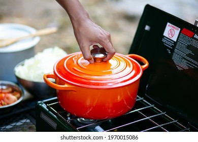 Man's Hand Putting Cover On A Red Enameled Cast Iron Dutch Oven On Camping Stove