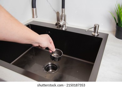 A man's hand removes a metal strainer from a kitchen sink drain. Cleaning the drain and pipes from clogging with food particles after washing dishes