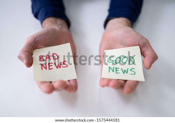 Man\'s Hand Showing Paper With Good And Bad News On\
White Desk