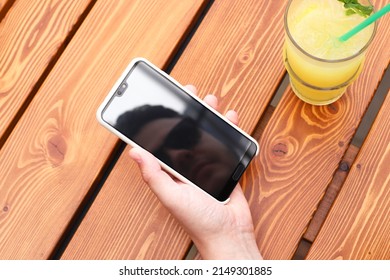 Man's Hand, Smartphone With Young Man's Face Reflection And Lemon Cocktail On Wooden Table. Top View