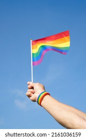 Man's Hand Wears LGBT Rainbow Wristband Is Waving LGBT Rainbow Flag On Background Blue Sky(selective Focus), Concept For LGBTQ+ Equality Movement Community