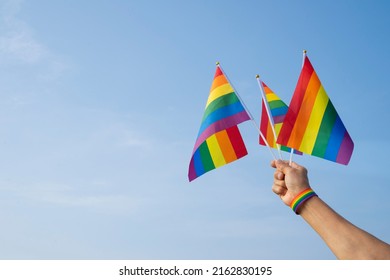 Man's Hand Wears LGBT Rainbow Wristband Is Waving LGBT Rainbow Flags On Background Blue Sky, Concept For LGBTQ+ Equality Movement Community