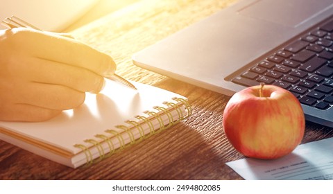 man's hand writes down information from a laptop in a notebook, next to a snack in the form of a fruit-an apple