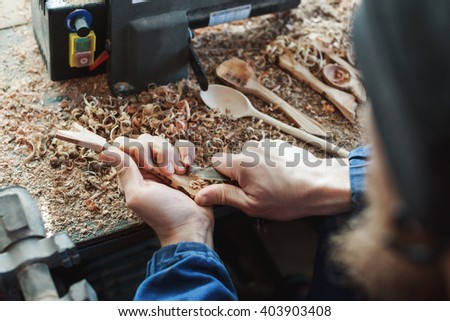 Man's hands in blue jeans working suit carving a wooden spoon with a knife, shavings on table at background, close up, woodworking, copy space.