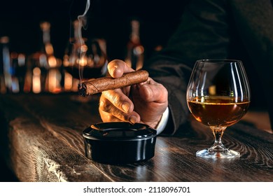 Man's hands with a cigar, elegant glass of brandy on the bar counter. Alcoholic drinks, cognac, whiskey, port, brandy, rum, scotch, bourbon. Vintage wooden table in a pub at night.