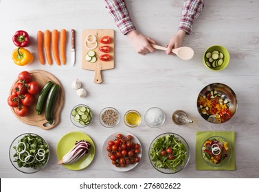 Man's Hands Cooking At Home Holding A Wooden Spoon, Fresh Vegetables And Food Ingredients All Around, Top View