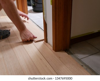 A Man's Hands Fitting The Final Hardwood Shining Gum (Eucalyptus Nitens) Floor Board In The Hallway Of A House Renovation, With White Walls And Grey Tiles Contrasting The Wood In Australia