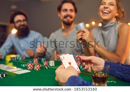 Mans hands holding playing cards with aces, poker chips and drink in glass during poker gambling over smiling friends faces at background, close-up, selective focus. Gambling, casino, poker concept