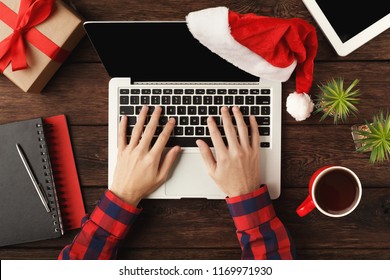 Man's hands on laptop. Work on Christmas Eve. Top view on human hands, computer keyboard, coffee, Santa hat, gift and digital tablet wooden table background. - Powered by Shutterstock