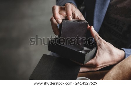 The man's hands take out a dark brown leather wallet from a black gift box. An elegant accessory symbolizes style, efficiency and attention to detail