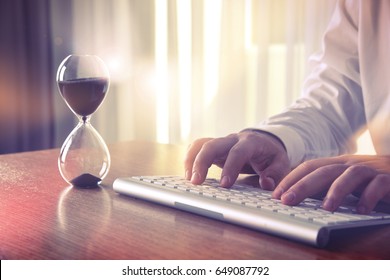 Man's hands typing on computer keyboard next to a hourglass. Concept of time management, business schedule and deadline, for background, website banner, promotional materials, advertising.