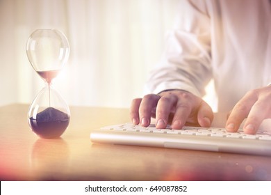 Man's Hands Typing On Computer Keyboard Next To A Hourglass. Concept Of Time Management, Business Schedule And Deadline, For Background, Website Banner, Promotional Materials, Advertising.