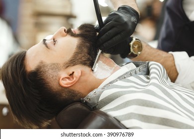 Man's Hands Wearing Black Gloves And Watch Making A Beard Form With Razor For Man With Dark Hair And Beard At Barber Shop, Portrait, Close Up.