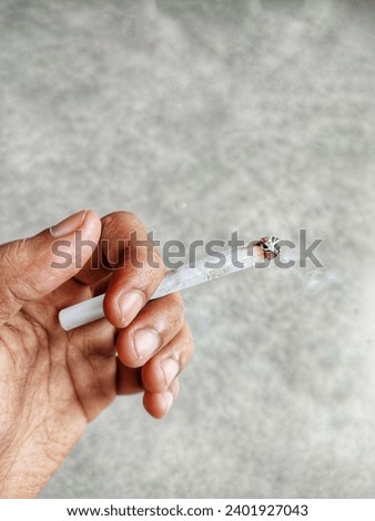 A man's left hand is holding a white lit cigarette