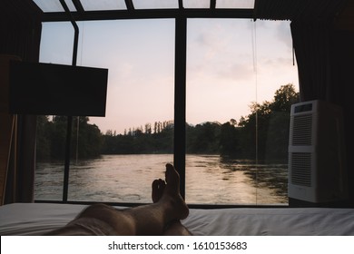 Man's Leg Lay On The Bed In The Room That Can See Through The River In Sunset Time With Silhouette Tone.