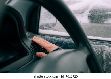 Man's Light-skinned Hand Is Warming Near A Car Heater In The Black Interior Of The Car Against The Background Of A Window In The Snow. Warming Up The Car In Bad Weather In Winter.