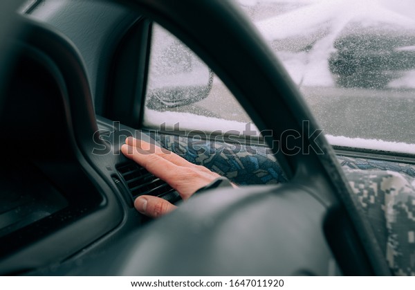 man\'s\
light-skinned hand is warming near a car heater in the black\
interior of the car against the background of a window in the snow.\
Warming up the car in bad weather in\
winter.