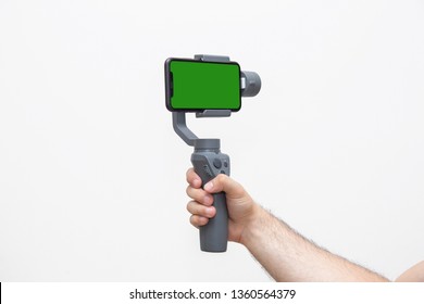 Man's right hand holding a gimbal stabilizer with mobile phone with green screen in  horizontal mode in a white background - Shutterstock ID 1360564379