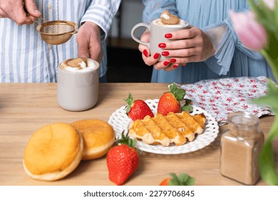 A mans and a woman's hands in light blue clothes in the background. A man ads cinnamon in a gray cup with caffe latte. The crispy waffle, red strawberries on a white plate and donuts. - Shutterstock ID 2279706845