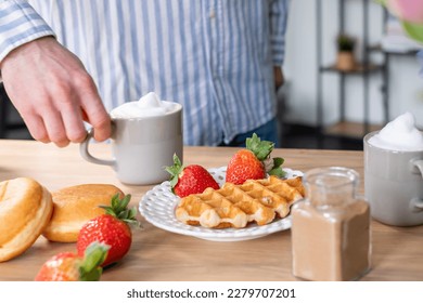 A mans and a woman's hands in light blue clothes with red nails. A man ads cinnamon in a gray cup with caffe latte. The crispy waffle, red strawberries on a white plate and donuts. - Shutterstock ID 2279707201