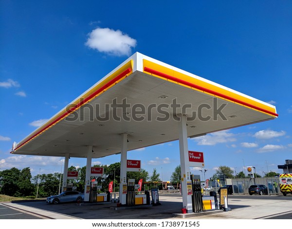 MANSFIELD, UK - MAY 9, 2020: Shell petrol\
station forecourt, 4 V-Power pumps, a car refueling under a large\
steel fabricated canopy. Cars and a van being clean in background,\
during COVID-19\
Pandemic
