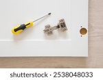 Manual screwdriver and hinge on white wardrobe door on floor background. Assembling new wooden furniture. Closeup. Top view.