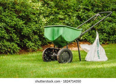 Manual walk behind grass seed spreader and bag of lawn fertilizer in a green residential backyard.
