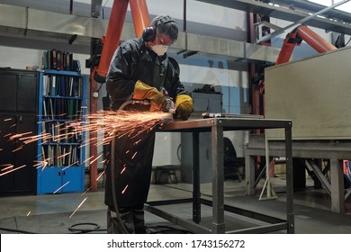 Manual Worker In Mask And Ear Protectors Standing At High Metal Table And Cutting Metal With Rotary Tool In Industrial Shop