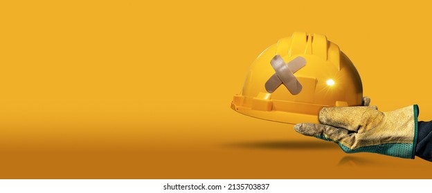 Manual worker with protective work glove holding a yellow safety helmet with a cross bandaid (adhesive bandage), on a yellow and orange background with copy space and reflections. Workplace security. - Shutterstock ID 2135703837