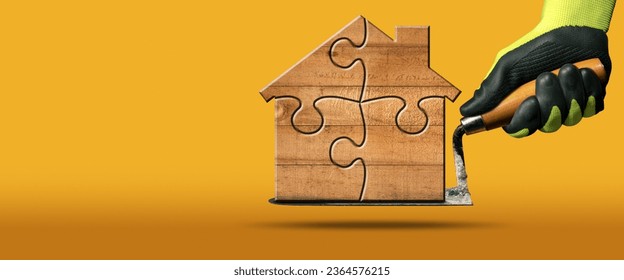 Manual worker with protective work gloves holding a dirty trowel and a small wooden house formed from jigsaw puzzle pieces. On yellow and orange background with shadow and copy space, photography. - Shutterstock ID 2364576215