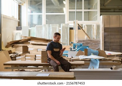 Manual Worker Sitting On Table And Using His Mobile Phone During His Break After Work In Warehouse