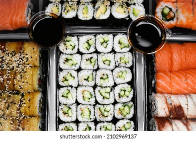 Many Assorted Fresh Fish And Vegan Rolls In Plastic Black Boxes On A Light Background With Jars Of Dark Soy Sauce