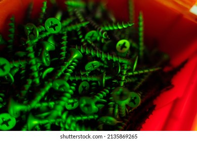 Many black self-tapping screws in a red container under green light, close-up