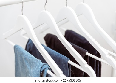 Many Blue Denim Jeans Hanging On White Clothes Hangers On Clothing Rack. Close Up Of Folded Casual Denim Jeans In Wardrobe With Coat Hangers